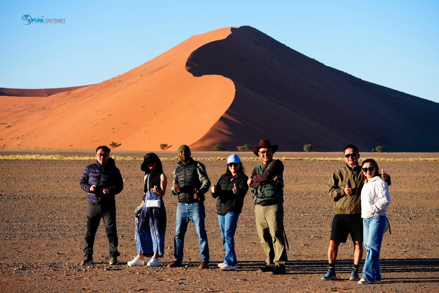 Group of tourists posing and giving thumbs up at the desert’s edge, with a tall orange sand dune and clear blue sky behind them.